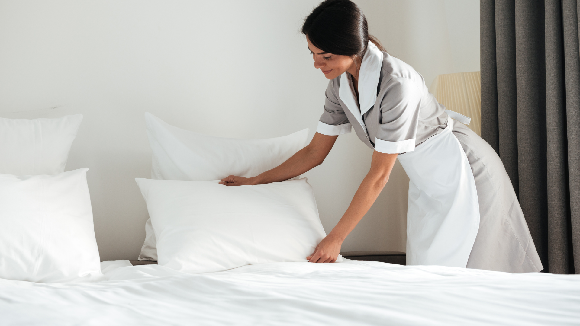 Femme de chambre disposant un oreiller sur un lit fraîchement préparé dans une chambre lumineuse.